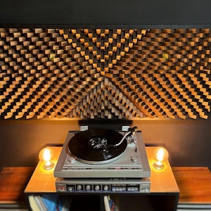 May include: A silver turntable with a black vinyl record sits on a wooden table, illuminated by two warm-toned light bulbs. Above, a large, geometric wooden wall art piece adds a decorative element to the room.