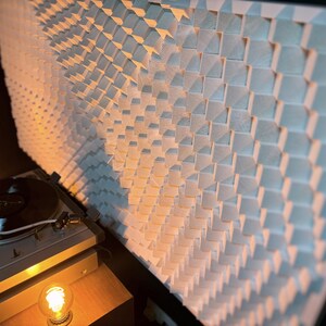 May include: A modern turntable with a black vinyl record playing, positioned in front of a large, textured wall panel made of light-colored wooden blocks. The scene is illuminated by warm, ambient lighting.