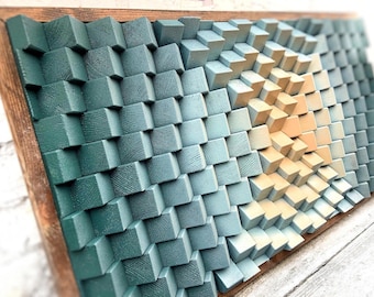 Op de afbeelding: Een wandkunstwerk gemaakt van houten blokken die in tinten blauw en beige zijn geverfd. De blokken zijn in een verspringend patroon gerangschikt, waardoor een 3D-effect ontstaat. Het stuk is ingelijst in hout.