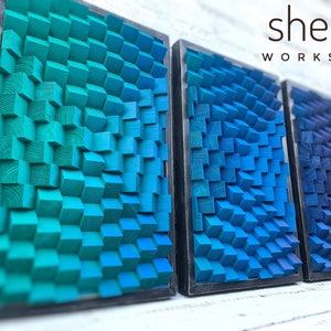May include: Three framed wall art pieces made of wood blocks. The blocks are arranged in a wave pattern and are painted in shades of blue and green. The frames are black and the pieces are hanging on a white brick wall. The text "shepit WORKSHOP" is visible in the upper right corner of the image.