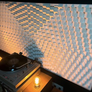 May include: A gray turntable with a black vinyl record sits on a wooden surface, illuminated by warm light. Behind it, a large white wall panel features a geometric pattern of small, angled squares, creating a textured effect.