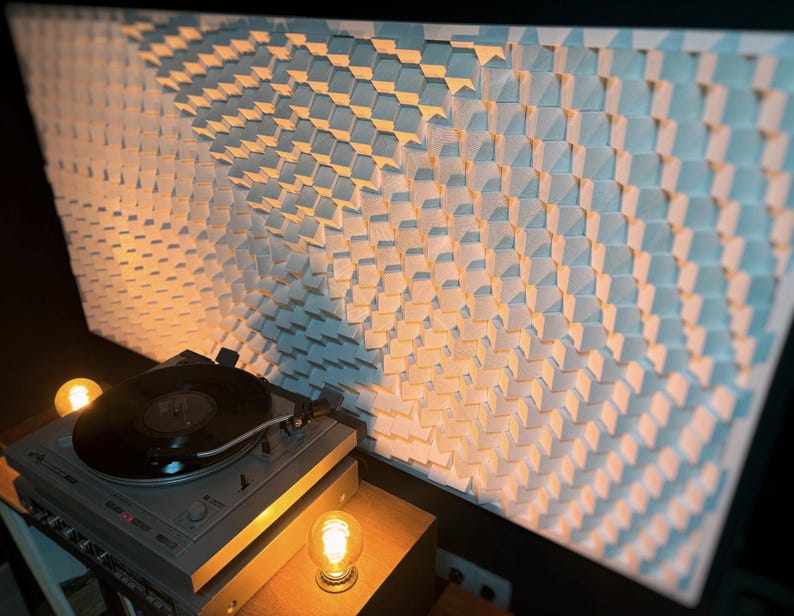 May include: A gray turntable with a black vinyl record sits on a wooden surface, illuminated by warm light. Behind it, a large white wall panel features a geometric pattern of small, angled squares, creating a textured effect.