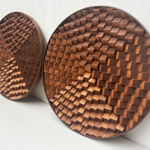 Puede incluir: Dos esculturas de pared redondas de madera con un patrón geométrico 3D. Las esculturas están hechas de pequeños bloques de madera cuadrados que están dispuestos en un patrón repetitivo. Las esculturas son de color marrón y tienen un acabado de madera natural.