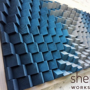 Può includere: Un'opera d'arte da parete realizzata con blocchi di legno in tonalità di blu e grigio, disposti a forma di onda. I blocchi sono impilati in modo sfalsato, creando un effetto 3D. Il pezzo è incorniciato in una cornice di legno naturale. Il testo "shepit WORKSHOP" è visibile nell'angolo in basso a destra.