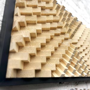 May include: A geometric wooden wall art piece with a black frame. The artwork features a repeating pattern of small, light-colored wooden blocks arranged in a stepped, wave-like design. The blocks create a textured, three-dimensional effect.