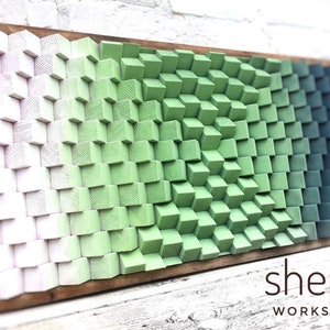 Può includere: Un'opera d'arte murale in legno con un gradiente di colori dal bianco al verde al blu. Il pezzo è composto da piccoli blocchi quadrati disposti in uno schema sfalsato. Il pezzo è incorniciato da una cornice in legno scuro. Il testo "shepit WORKSHOP" è visibile nell'angolo in basso a destra dell'immagine.