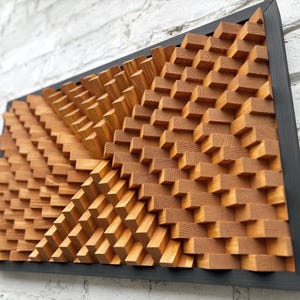 May include: A wooden wall art piece with a geometric design. The artwork features a series of small, rectangular blocks arranged in a repeating pattern, creating a textured surface. The blocks are a warm, natural wood tone, set within a black frame.