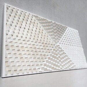 May include: A white, geometric wooden wall art piece. The artwork features a repeating pattern of small, square blocks arranged in a textured, three-dimensional design. The blocks are painted white and mounted on a white frame.