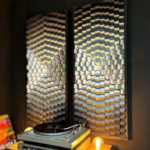 May include: Two rectangular wooden acoustic panels with a 3D cube design in shades of grey and natural wood tones. A vintage turntable sits below the panels, with a record on the platter. A warm light bulb illuminates the scene.