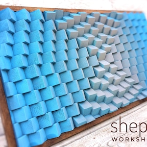 May include: A wall art piece made of wooden blocks painted in shades of blue and grey. The blocks are arranged in a geometric pattern, creating a 3D effect. The piece is framed in a dark wood frame. The text "shepit WORKSHOP" is visible in the bottom right corner of the image.