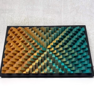 May include: A rectangular wooden wall art piece with a black frame. The artwork features a geometric design with blocks in shades of yellow, orange, and teal. The blocks are arranged in a pattern that creates a three-dimensional effect.