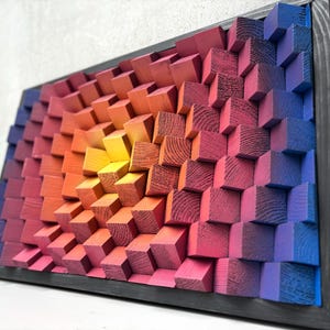 Op de afbeelding: Een houten kunstwerk in een frame met een kleurengradiënt. Het stuk bestaat uit veel kleine, vierkante blokken, waardoor een gestructureerd, driedimensionaal effect ontstaat. Kleuren gaan over van geel naar oranje, roze en blauw.