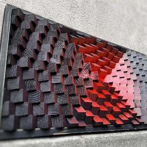 Könnte beinhalten: Ein Wandkunstwerk aus Holzblöcken, die in Schwarz- und Rottönen bemalt sind. Die Blöcke sind in einem geometrischen Muster angeordnet, das einen 3D-Effekt erzeugt. Das Stück ist in Schwarz gerahmt.