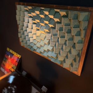 May include: A wall art piece made of wood blocks in shades of blue and green, arranged in a geometric pattern. The blocks are painted with a wood grain texture, giving the piece a rustic look. The piece is framed in a natural wood frame.