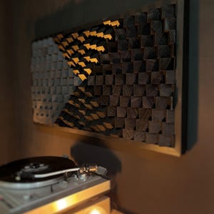 May include: A wooden wall art piece with a geometric design. The artwork features a pattern of small, square wooden blocks in shades of gray, black, and natural wood tones, arranged in a rectangular frame. The artwork is mounted on a brown wall, with a record player in the foreground.