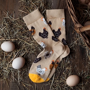 Puede incluir: Un par de calcetines color beige con un estampado repetido de gallinas de varios colores. Los calcetines tienen talones amarillos y se exhiben sobre un lecho de heno con tres huevos blancos y una cesta de huevos en el fondo.