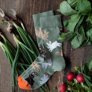 May include: A pair of green socks with a repeating pattern of running rabbits in white, brown, gray, and black. The socks have orange toes and are displayed on a wooden surface with green onions and radishes.