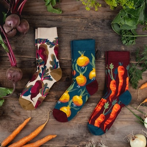 Puede incluir: Tres pares de calcetines con estampados de verduras. El primer par es crema con remolachas, el segundo es azul verdoso con cebollas amarillas y el tercero es rojo oscuro con zanahorias naranjas. Cada calcetín tiene el talón y la puntera en contraste.