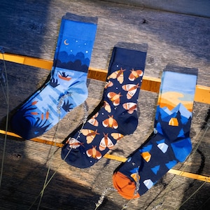 May include: Three pairs of crew socks featuring distinct outdoor designs. The left sock shows a night lake scene with a boat, the middle sock displays a pattern of moths on a dark background, and the right sock depicts a mountain landscape with a tent.