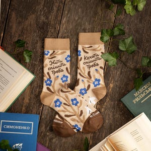 Puede incluir: Un par de calcetines beige con flores azules y pájaros blancos. Los calcetines tienen el texto "Жити спишити Треба" y "Кохати спишити Треба" impreso en ellos.