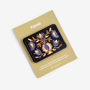 Könnte beinhalten: Ein schwarzer, bestickter Aufnäher mit einem Blumenmuster in Blau, Weiß und Orange, der auf einer beigen Karte liegt. Das Wort "Крим" ist in Weiß auf die Karte gedruckt. Der Aufnäher hat ein detailliertes, farbenfrohes Blumenmuster.