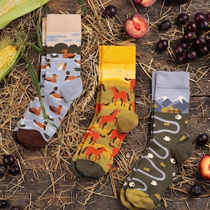 May include: Three pairs of crew socks featuring distinct patterns: one light blue pair with ducks on a pond, one yellow pair with red horses in a field, and one olive green pair with white sheep grazing in a landscape with mountains.