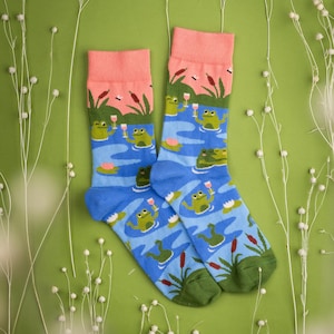 Puede incluir: Un par de calcetines azules y rosas con un diseño caprichoso de ranas verdes bebiendo vino en un estanque. Los calcetines tienen un puño verde y están rodeados de flores blancas.