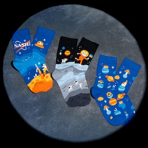 May include: Three pairs of crew socks with space-themed designs. One pair is blue with the text "NASHI" and a rocket ship. Another pair is black and grey with an astronaut. The third pair is blue with planets and mushrooms.