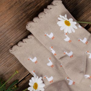 May include: A pair of beige socks featuring a repeating pattern of small white geese with orange beaks and feet. The socks have a scalloped, ruffled edge at the top opening. Two white daisy flowers with yellow centres are placed near the socks.