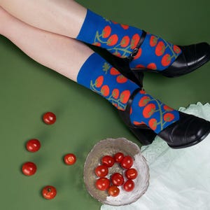 以下が含まれることがあります： 赤いトマトと緑のつるの模様が入った青い靴下。黒い靴を履いています。銀色のボウルとバラバラのトマトが緑色の表面に置かれています。