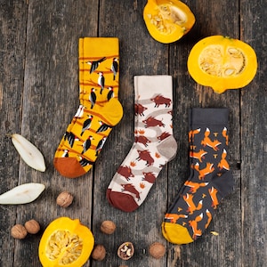 May include: Three pairs of crew socks featuring animal patterns. One pair is yellow with black and white woodpeckers on branches, one is beige with brown wild boars, and one is dark gray with orange foxes. Each sock has a solid colored toe and heel.