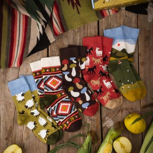 May include: A selection of patterned socks featuring sheep, mushrooms, horses, and geometric designs. The socks are displayed on a wooden surface alongside apples, corn, and a decorative box. The socks are in various colours.