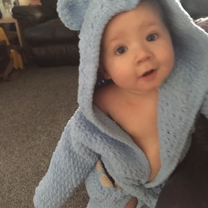 May include: A baby wearing a light blue hooded robe with bear ears. The robe is made of a soft, fuzzy material and has a tie at the waist.