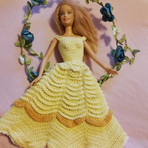 Puede incluir: Una muñeca con un vestido amarillo de ganchillo hecho a mano con un dobladillo festoneado y detalles melocotón. La muñeca tiene el pelo rubio y está colocada frente a una corona floral con flores azules y hojas verdes. El fondo es rosa claro.