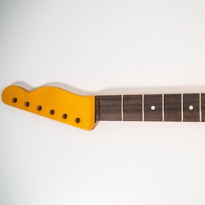 May include: A dark brown guitar neck with a light brown headstock and white dot inlays. The neck is unfinished and ready to be assembled.