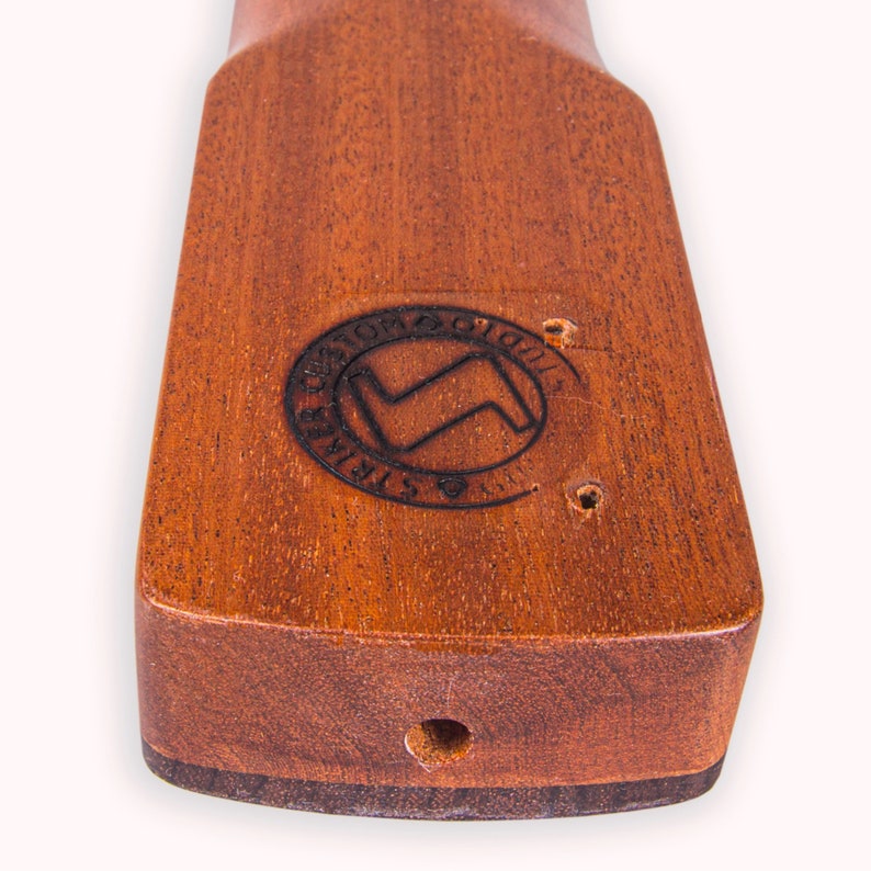 May include: Close-up of a wooden ukulele neck with a dark brown stain. The neck has a circular logo that reads "Custom Guitars" with a lightning bolt symbol inside.