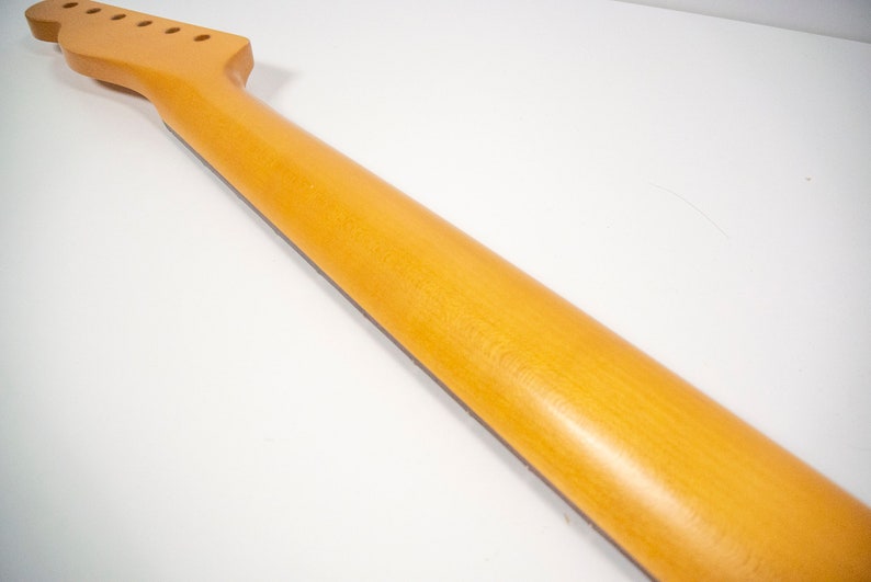 May include: A natural wood guitar neck with a light brown finish. The neck has six tuning pegs and a fretboard.