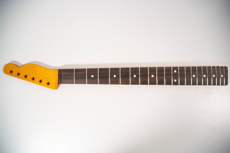 May include: A dark brown guitar neck with a light brown headstock and white dot inlays. The neck is unfinished and ready to be assembled.