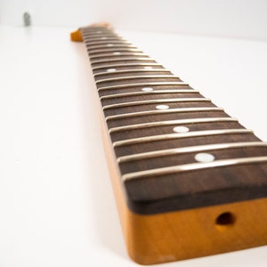 May include: A close-up of a guitar neck with a rosewood fretboard and white dot inlays. The neck is unfinished and has a natural wood color.
