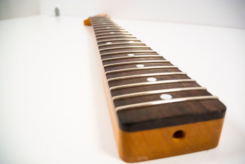 May include: A close-up of a guitar neck with a rosewood fretboard and white dot inlays. The neck is unfinished and has a natural wood color.