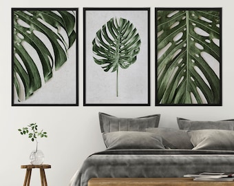 May include: Three framed botanical prints featuring close-up images of green Monstera leaves. The prints are displayed above a bed with gray bedding and a wooden bench. A small wooden side table holds a glass vase with greenery.