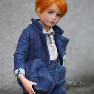 May include: A porcelain doll with red hair wearing a blue suit and holding a blue hat. The doll has a white shirt with a bow tie and a pair of braces. The doll is looking down and has a serious expression on its face. The doll is standing on a grey background. @juliartdolls