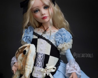 May include: A doll dressed in a light blue dress with white lace trim and puffed sleeves. The doll has blonde hair, blue eyes, and a black bow. She holds a stuffed rabbit and a small bottle. The doll's outfit includes a black and white checkered vest.