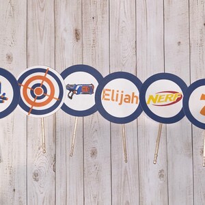May include: A collection of circular party decorations on wooden sticks. Each circle has a navy blue border and a white center with various designs, including a target, a toy gun, the name "Elijah", the word "NERF", and the number "7".