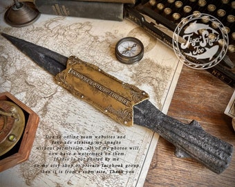 May include: A decorative dagger with a dark blade and a gold-colored hilt. The hilt features ornate detailing and text. The dagger rests on a vintage map, with a compass and other antique items in the background.