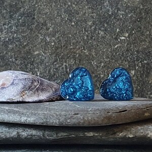 Puede incluir: Un par de pendientes de botón con forma de corazón. Los pendientes son de un azul vibrante con un aspecto texturizado y brillante. Se exhiben sobre una superficie de piedra gris, con una concha en el fondo.