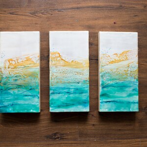 Può includere: Tre dipinti astratti su tela con uno schema di colori blu, verde e giallo. I dipinti sono disposti in fila e rappresentano un paesaggio marino con onde e un orizzonte dorato.