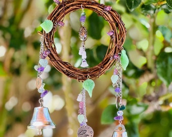 Puede incluir: Un carillón artesanal con un círculo de vid trenzada, adornado con cuentas moradas y verdes, detalles en forma de hojas y tres campanas plateadas. Cuelga de una cadena plateada, creando un ambiente caprichoso y decorativo.