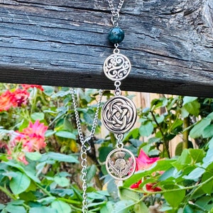 May include: A silver necklace with a chain and multiple pendants. The pendants include a green bead, a circular filigree design, a Celtic knot, and a thistle. The necklace is displayed against a backdrop of greenery and flowers.