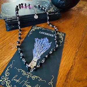 May include: A black beaded necklace with a silver clasp rests on a black card with the word "Lavender" and a lavender illustration. A stack of tarot cards and a lit candle in a black cauldron are in the background.
